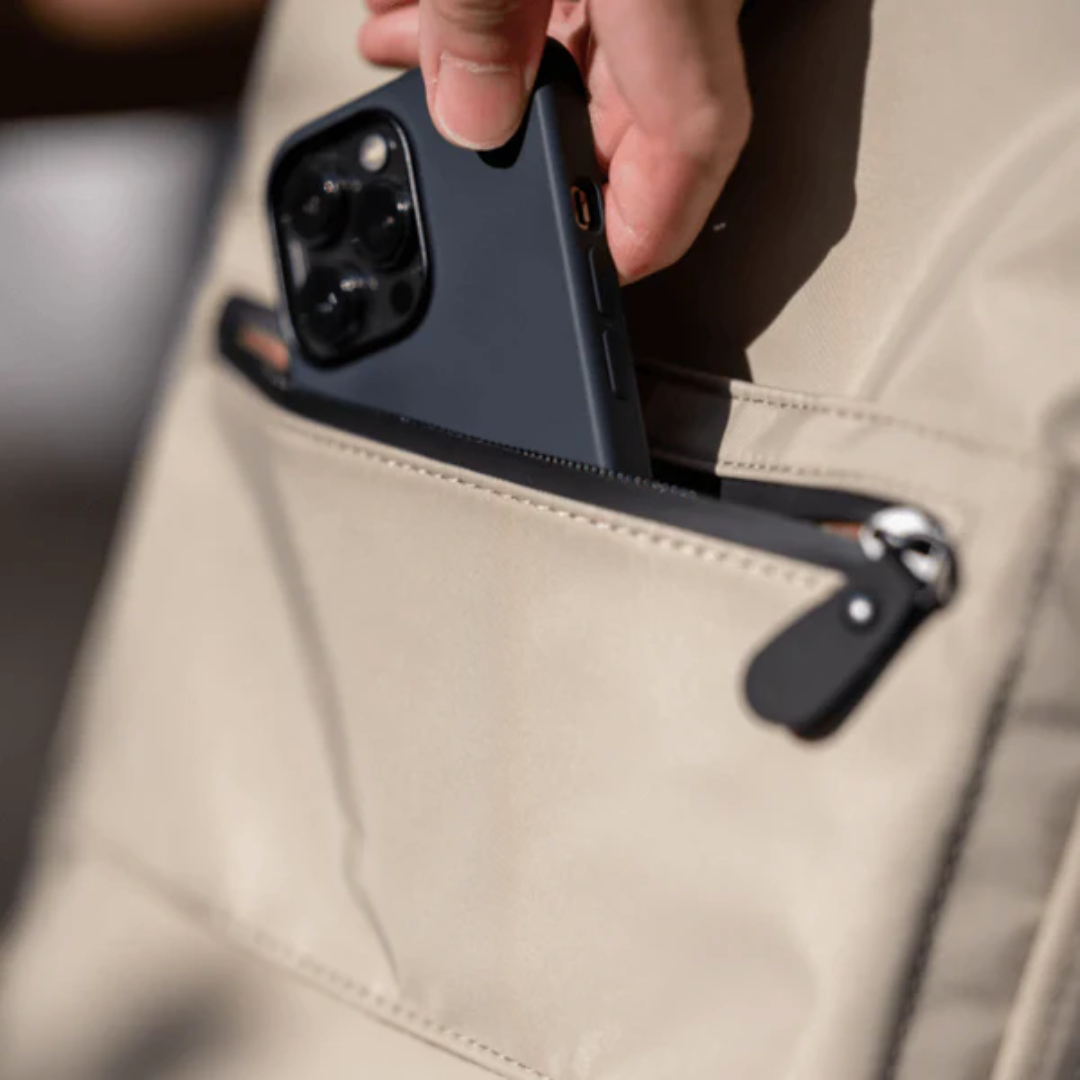 Hand placing a smartphone into the front zipper pocket of a beige backpack.