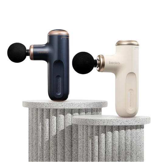 Beoka handheld massage guns in navy blue and ivory white, each with a round black massage head, displayed upright on textured stone pedestals against a white background