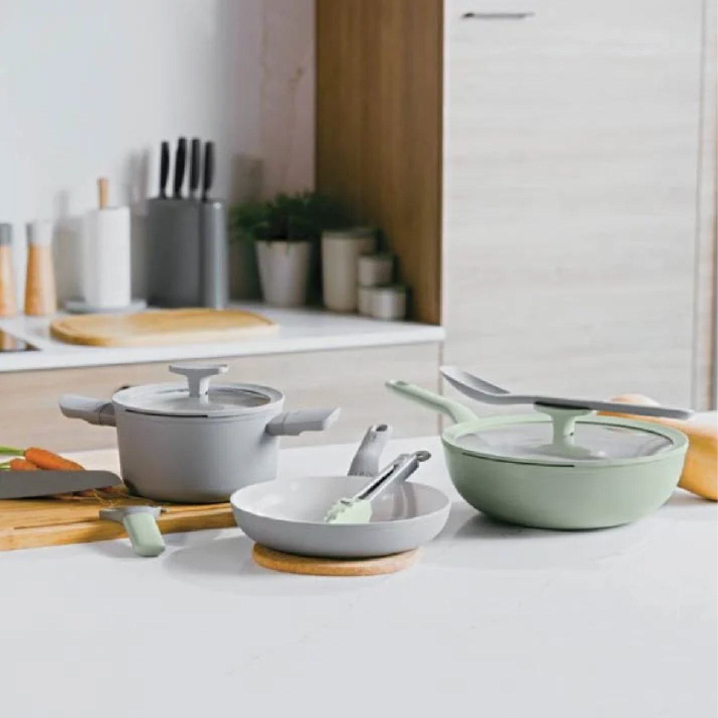 Modern kitchen scene featuring BergHOFF sage green and grey ceramic non-stick pots, pans and utensils arranged on a white countertop
