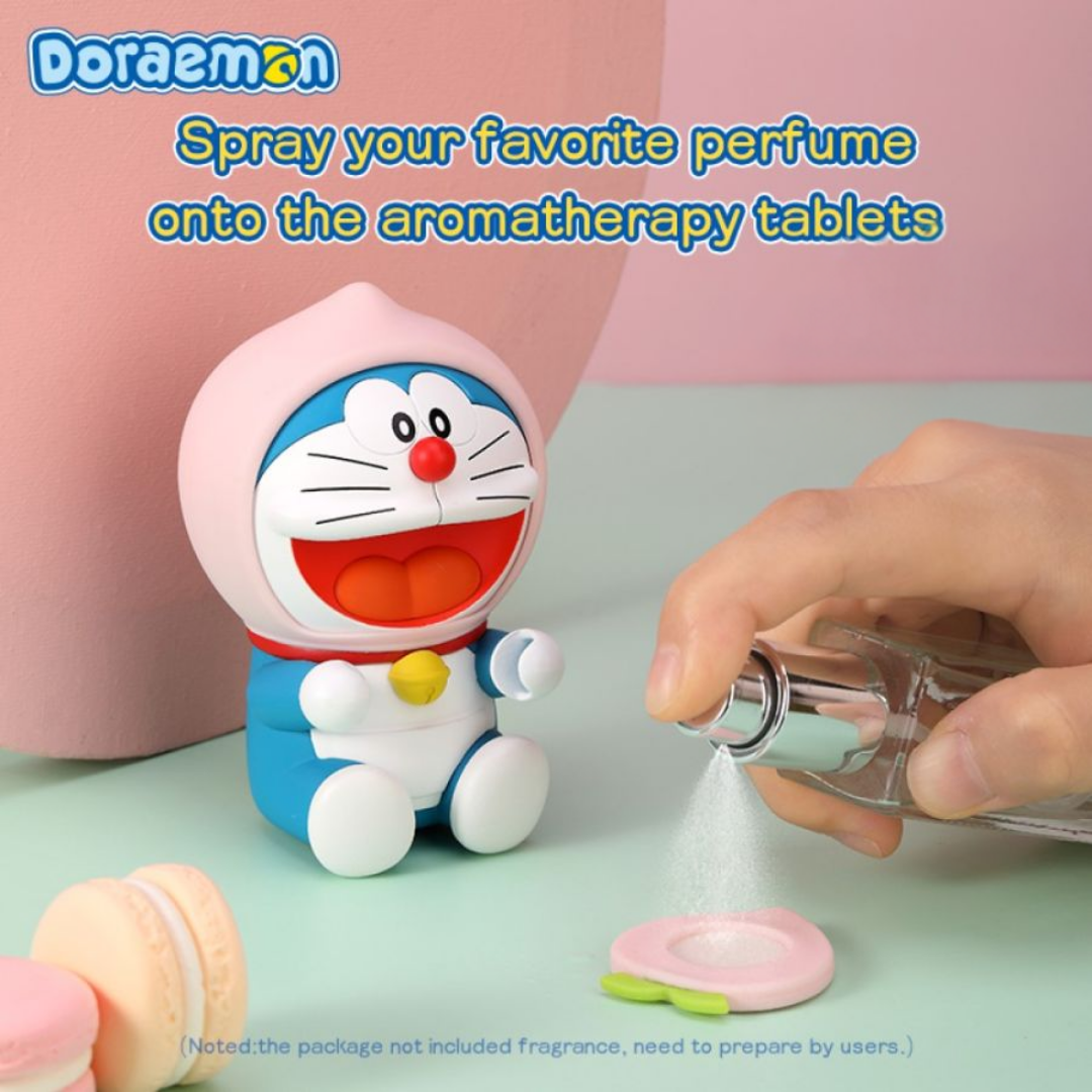 Hand spraying perfume onto a pink aromatherapy tablet in front of a Doraemon Fruit Aromatherapy diffuser shaped like a smiling Doraemon with a pink peach hood, on a pastel background with macarons and instructional text.