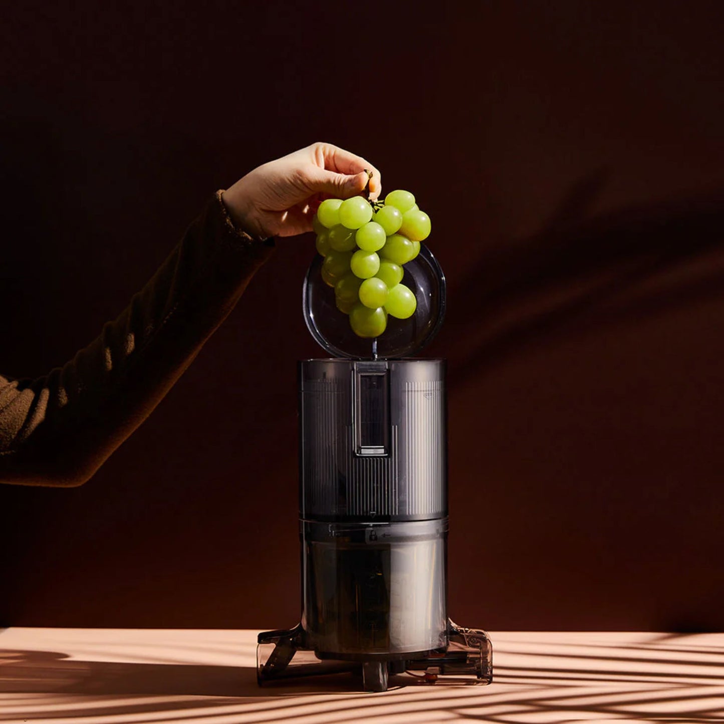 Hurom H310A Slow Juicer black model, BPA-free compact juicer with hand inserting green grapes into hopper, lifestyle action shot with dramatic lighting