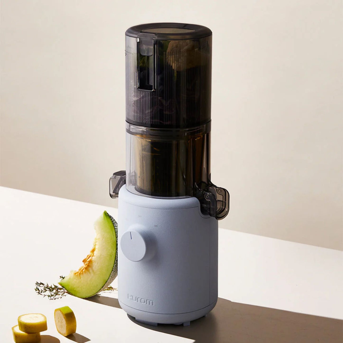 Hurom H310A Slow Juicer light blue model, BPA-free compact juicer shown in lifestyle setting with melon slice, banana slices, and herbs