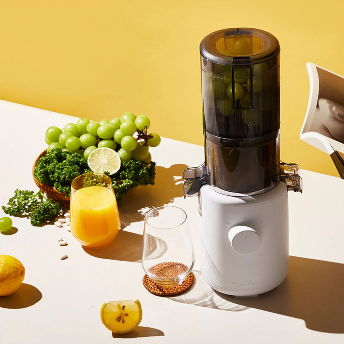 Hurom H310A Slow Juicer white model, BPA-free compact juicer in lifestyle scene with green grapes, kale, lemon, juice glass, and yellow background