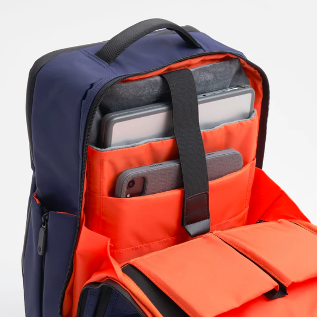Open navy backpack with bright orange interior, showing padded compartments securely holding a laptop, tablet, and smartphone.