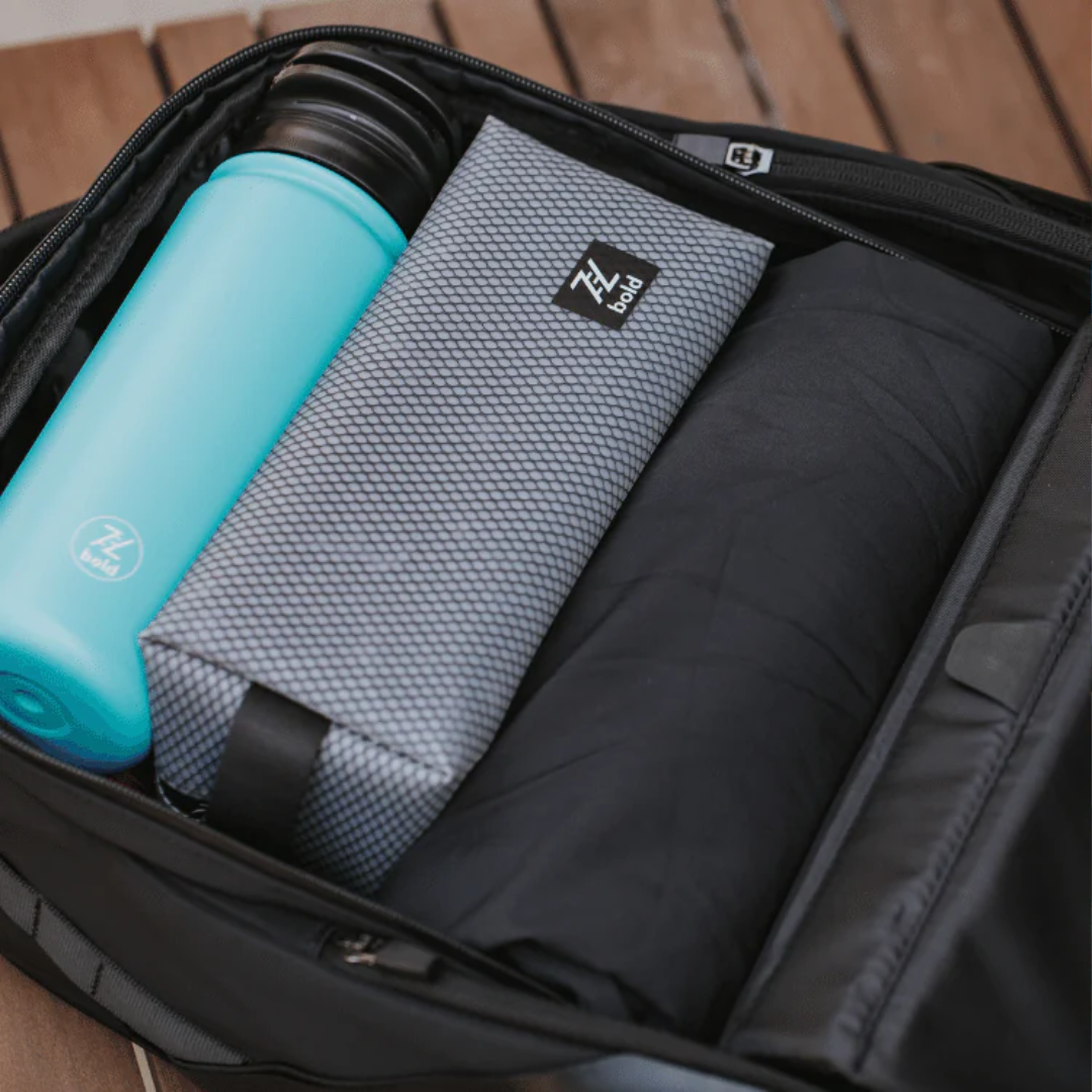 Top view of an open black backpack packed with a blue water bottle, a grey ZL bold pouch, and a rolled black jacket on a wooden surface.
