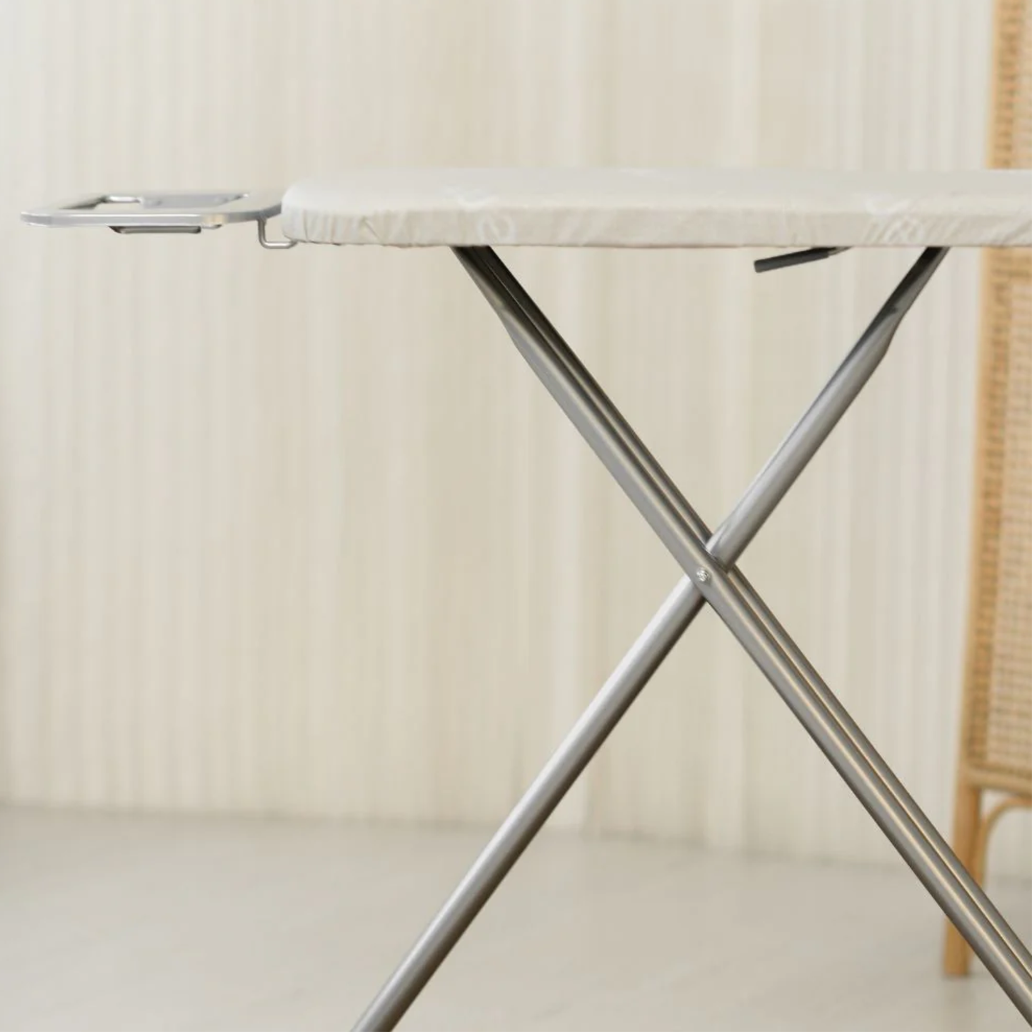 Close-up of RENE classic ironing board with padded white top and sturdy silver X-frame legs on a round jute rug beside a rattan room divider.