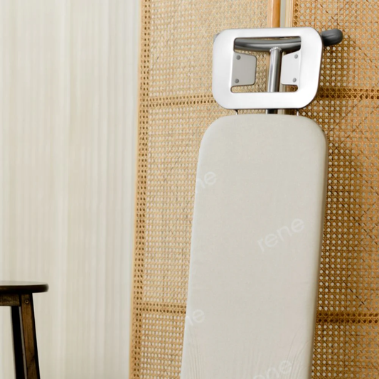 RENE classic ironing board with padded white cover and metal iron rest standing upright against a rattan room divider in a modern home.