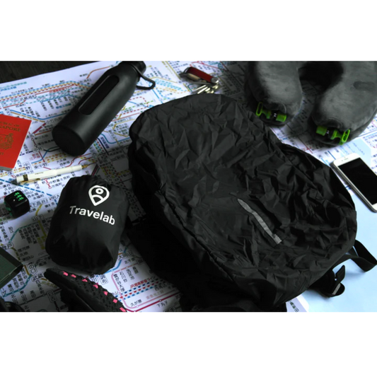 Travelab Freedom Pack black packable anti-theft backpack and compact storage pouch displayed with travel essentials including a water bottle, neck pillow, phone, and passport on a subway map background.