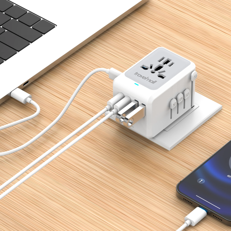 Travelmall universal multi-port charging adapter with multi-standard socket, sliding international plugs, USB-C and USB-A ports, plugged into a power strip to simultaneously charge a laptop and smartphone at a modern workstation.