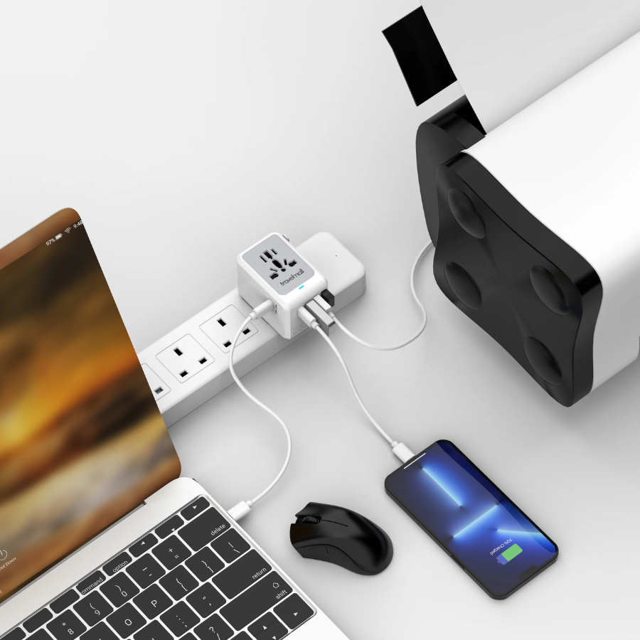 Travelmall universal multi-port power adapter plugged into a power strip, charging a laptop, smartphone, and printer with multiple USB and international plug ports at a modern workstation.