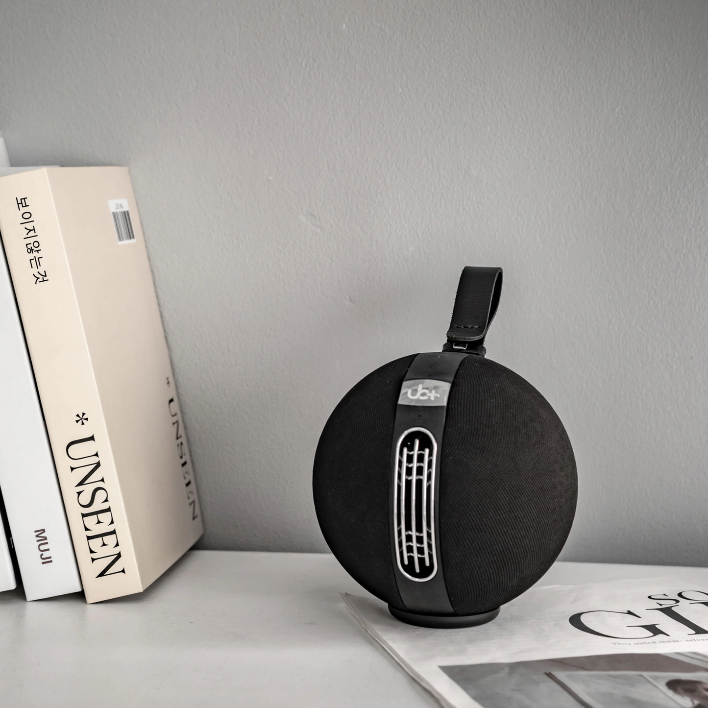 UB+ S1 Circle Bluetooth speaker in black, shown on a shelf beside minimalistic books and a magazine, with a leather strap and metallic panel detail.