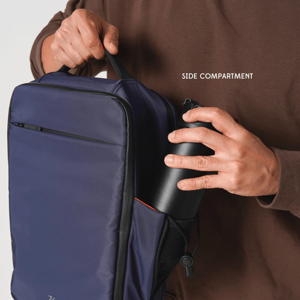 Bold Quiver 13L navy blue minimalist backpack with side compartment, man placing black water bottle into mesh pocket, top handle and front zipper pocket visible