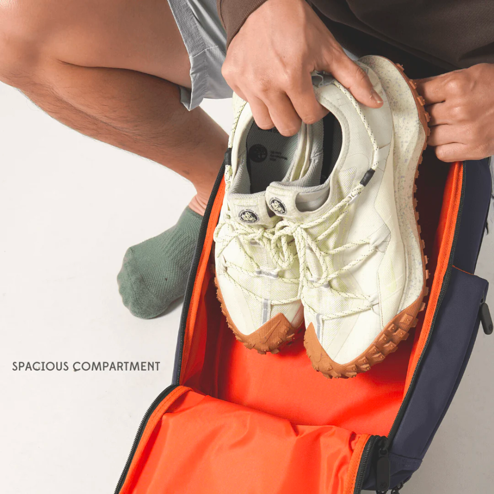 Bold Quiver 13L navy blue sports backpack with spacious bright orange shoe compartment, man placing white sneakers inside, ideal for gym, travel, and athletic organization
