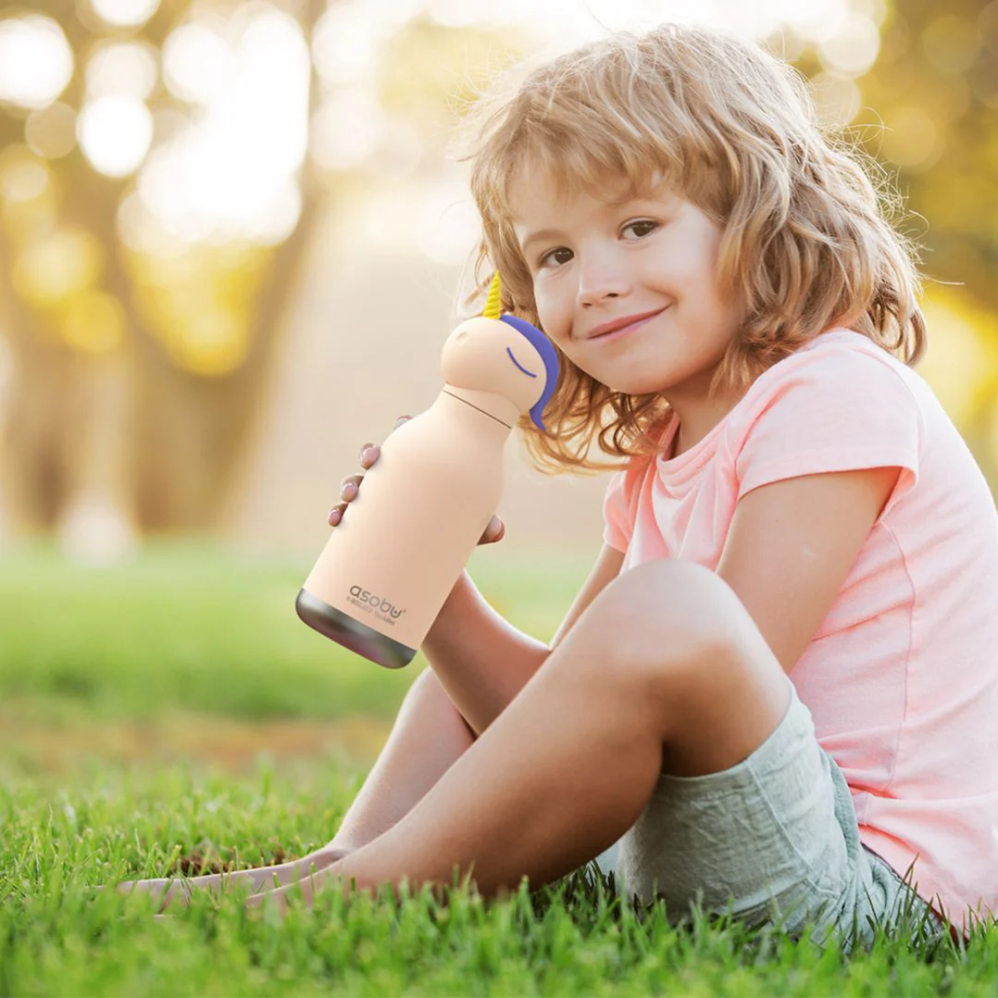 Smiling child sitting on grass, holding Asobu Bestie Bottle unicorn insulated water bottle, perfect for kids’ hydration and outdoor activities.