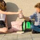 Two students sitting on pavement, sharing Asobu Bestie Bottle unicorn near backpacks, showcasing cute insulated water bottle for school.