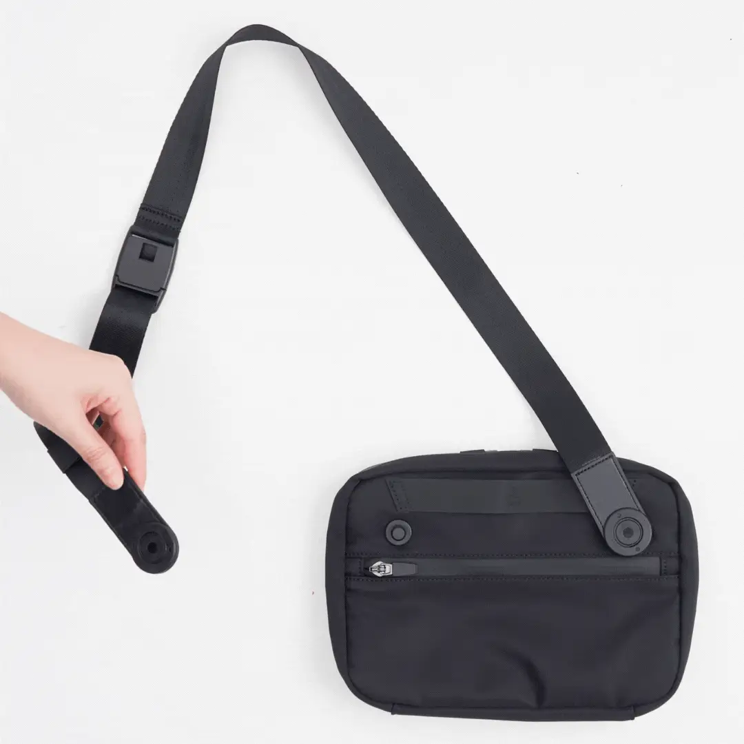 Hand adjusting the strap of a black crossbody sling bag with adjustable buckle and front zipper pocket on a white background.