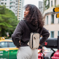 Bold Mimic Sling Bag in earth beige, worn crossbody on back by a person in an urban outdoor setting; compact style for city travel and daily carry.