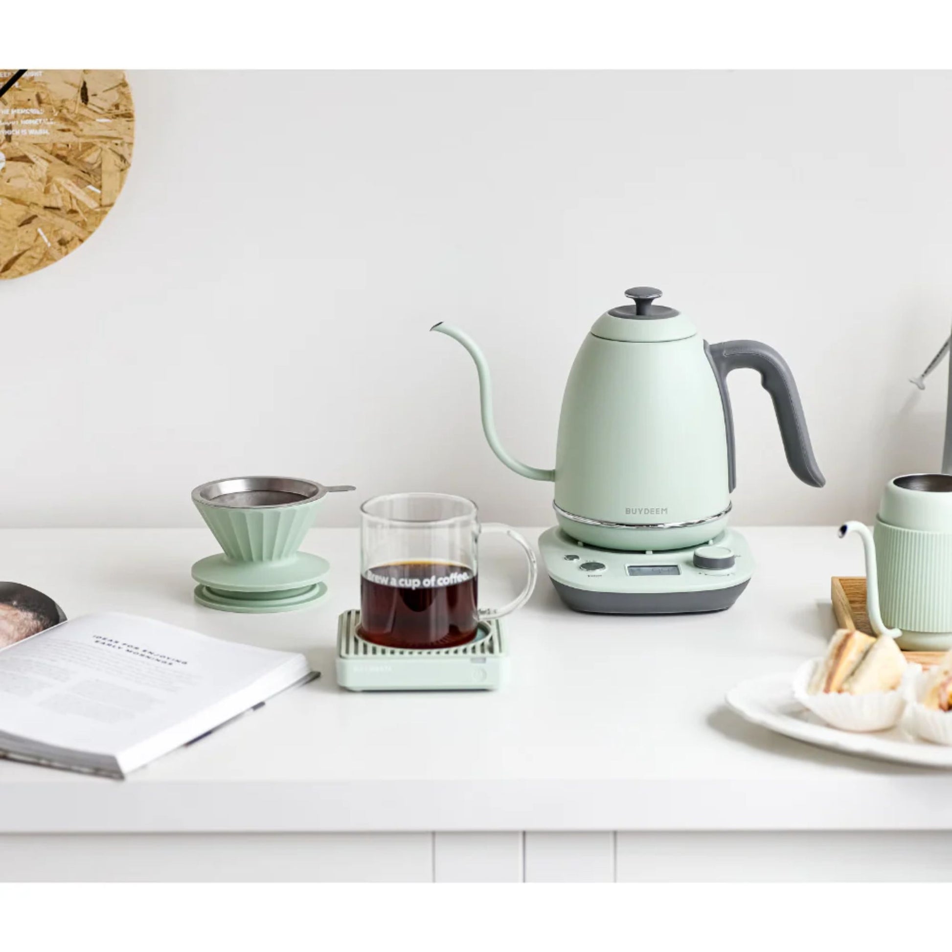 Buydeem mint green electric kettle with matching pour over coffee maker, glass mug, and pastries on a white kitchen countertop in a stylish modern breakfast scene