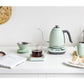 Buydeem mint green electric kettle with matching pour over coffee maker, glass mug, and pastries on a white kitchen countertop in a stylish modern breakfast scene