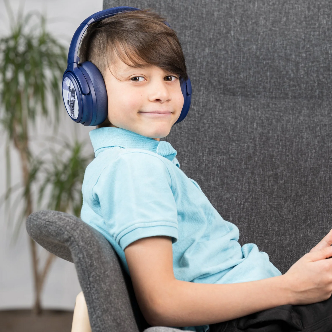 Child wearing blue over-ear headphones with a character design, sitting in a gray chair and holding a tablet, dressed in a light blue polo shirt; indoor setting with a plant in the background