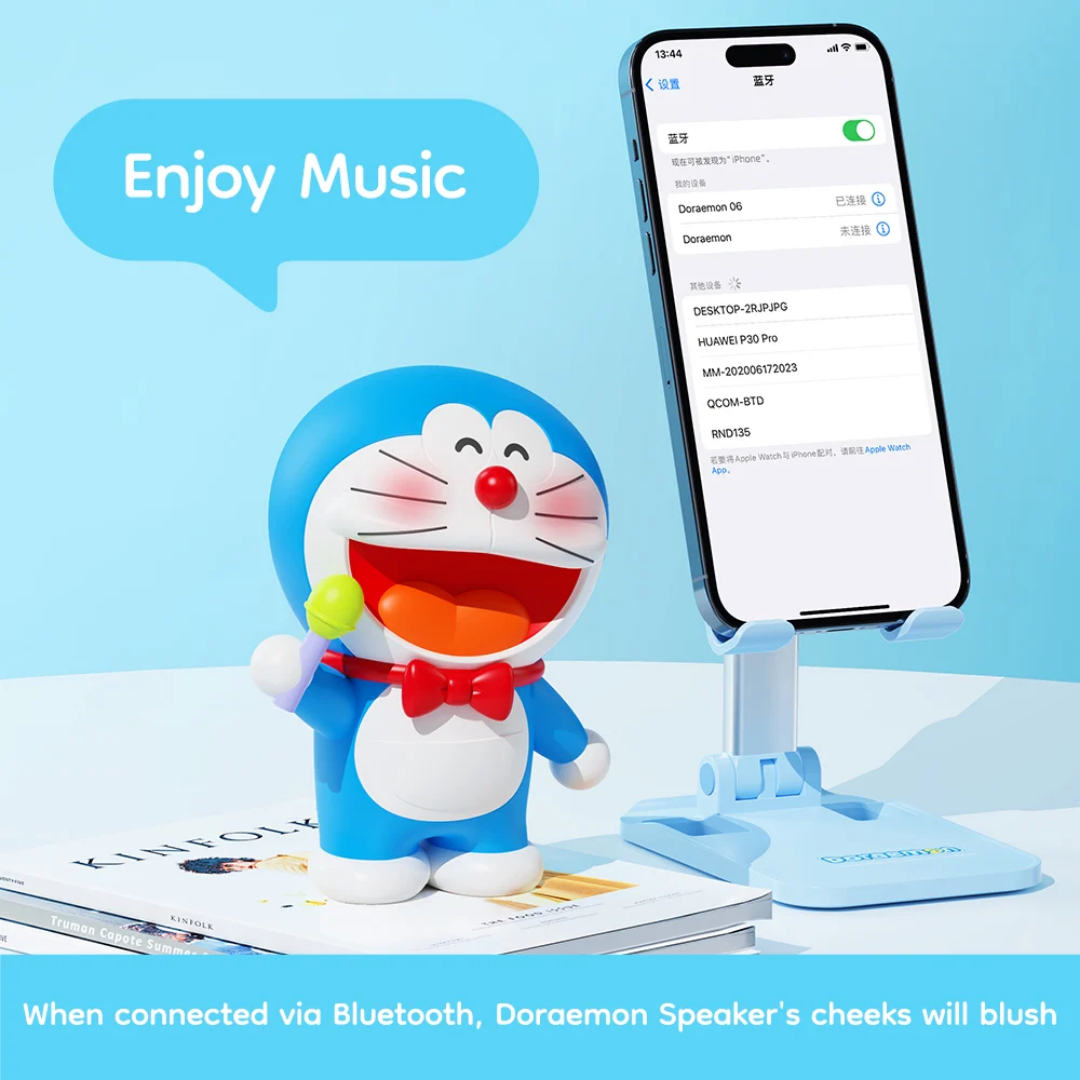 Doraemon Mic King Bluetooth Speaker with blushing cheeks, holding a green microphone, connected to a smartphone via Bluetooth, on a white desk with books and a blue phone stand, "Enjoy Music" text above.