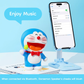 Doraemon Mic King Bluetooth Speaker with blushing cheeks, holding a green microphone, connected to a smartphone via Bluetooth, on a white desk with books and a blue phone stand, "Enjoy Music" text above.