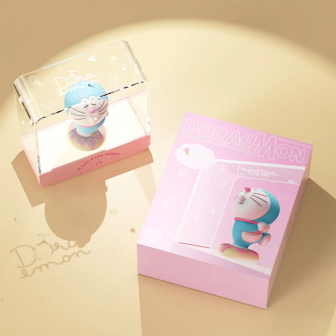 Doraemon Starry Night Light featuring a 3D Doraemon figure with pink nose and collar standing on a donut-shaped base inside a transparent rectangular lamp, displayed next to a matching pink Doraemon gift box on a beige surface with a Doraemon shadow.