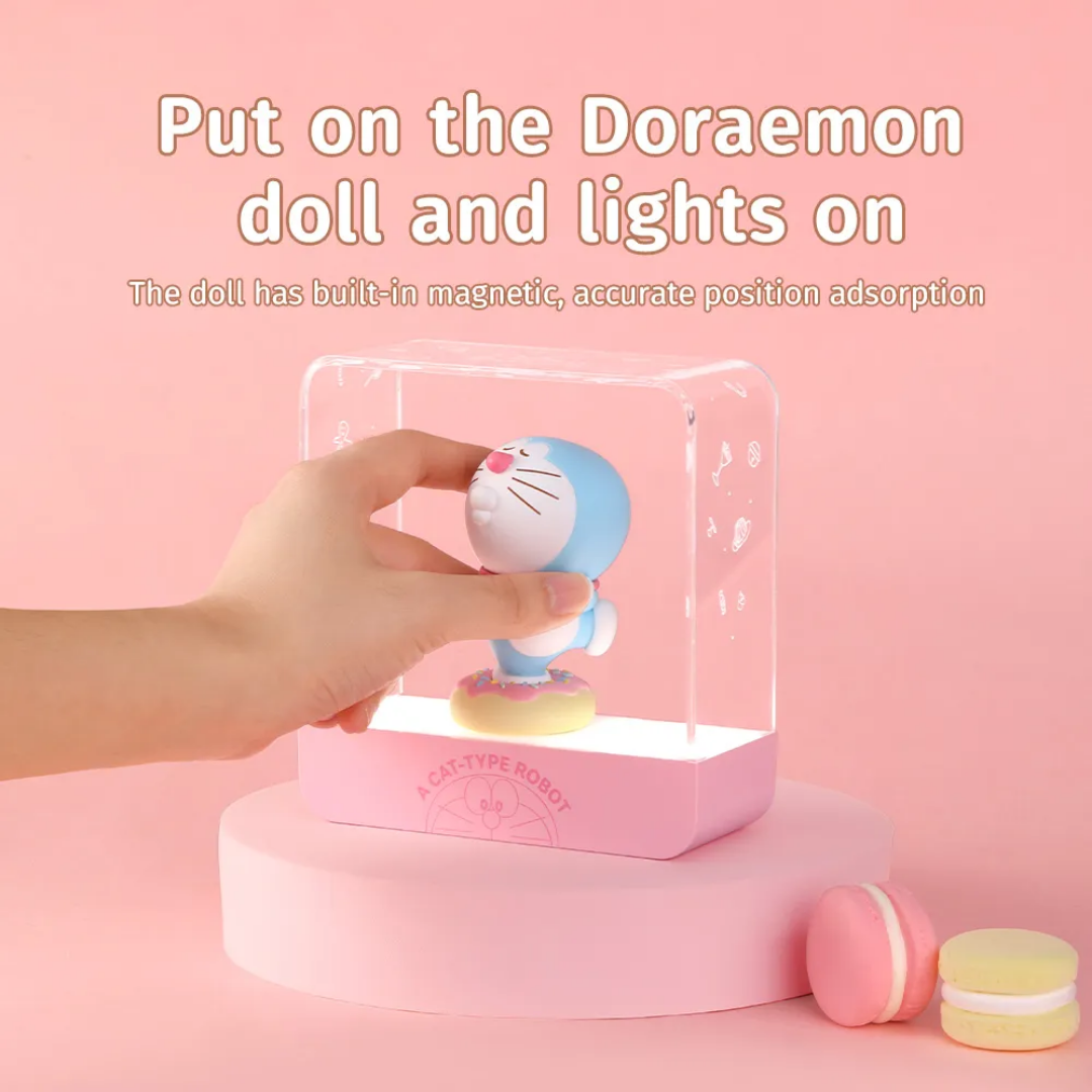 Hand placing a 3D Doraemon doll with built-in magnet onto a pink starry night light base, activating the soft LED glow, with macarons and "Put on the Doraemon doll and lights on" text on a pastel pink background