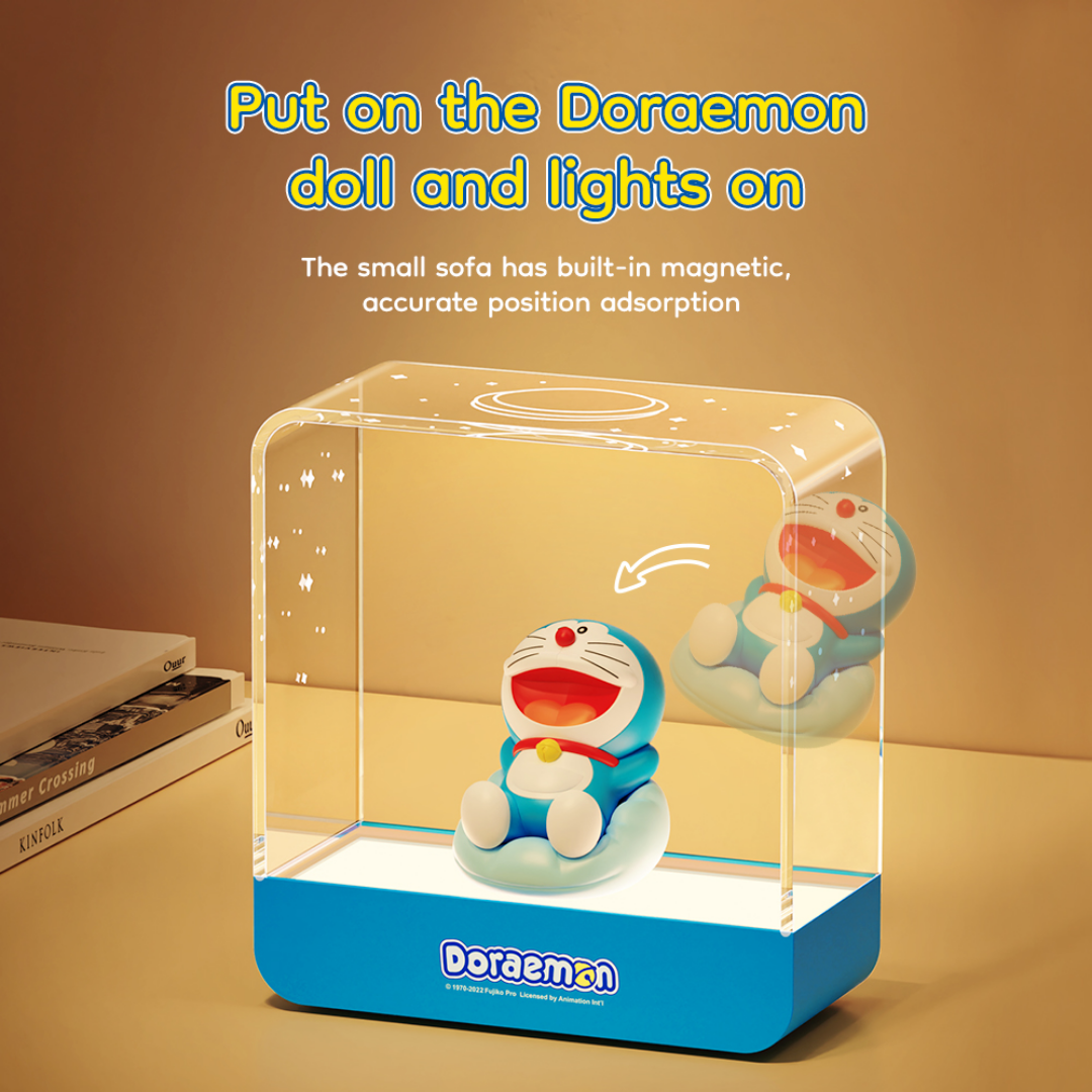 Doraemon Starry Night Light with magnetic sofa, featuring a smiling Doraemon doll seated inside a transparent rectangular lamp that illuminates when the doll is placed, on a bedside table with books