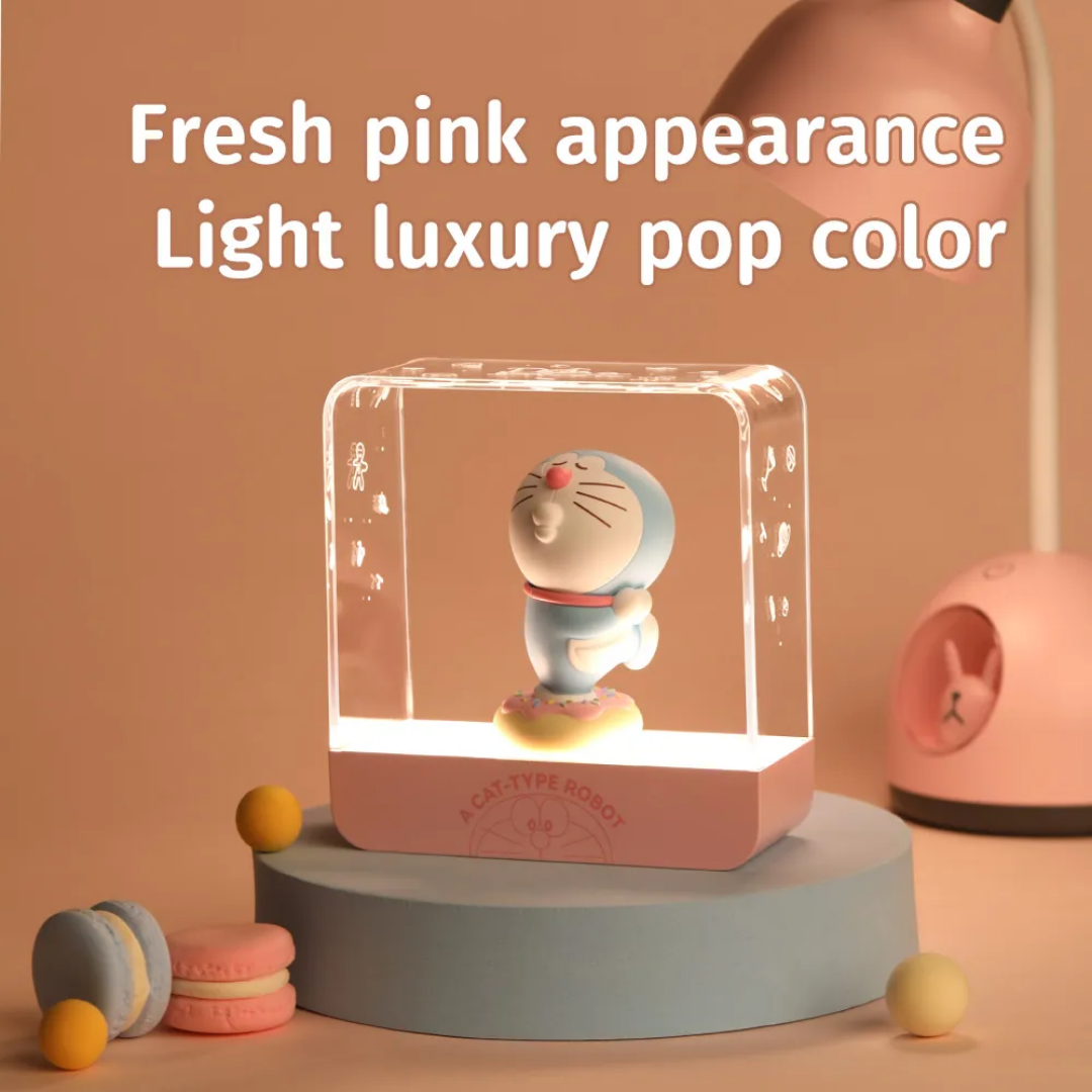 Doraemon Starry Night Light with a 3D Doraemon figure standing on a donut-shaped base inside a transparent rectangular lamp, featuring a fresh pink appearance and soft LED glow, displayed on a pastel podium with macarons and a pink desk lamp in the background.