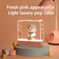 Doraemon Starry Night Light with a 3D Doraemon figure standing on a donut-shaped base inside a transparent rectangular lamp, featuring a fresh pink appearance and soft LED glow, displayed on a pastel podium with macarons and a pink desk lamp in the background.