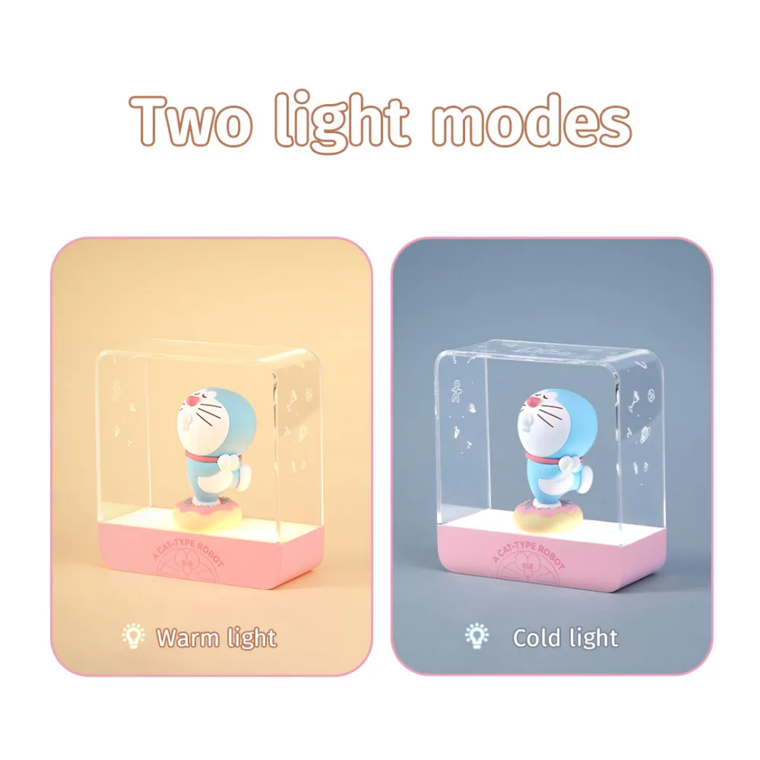 Doraemon Starry Night Light showing two lighting modes: warm light with a soft yellow glow and cold light with a cool white glow, each featuring a 3D Doraemon figure inside a transparent rectangular lamp with a pink base, on matching colored backgrounds. "Two light modes" text above