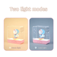 Doraemon Starry Night Light showing two lighting modes: warm light with a soft yellow glow and cold light with a cool white glow, each featuring a 3D Doraemon figure inside a transparent rectangular lamp with a pink base, on matching colored backgrounds. "Two light modes" text above