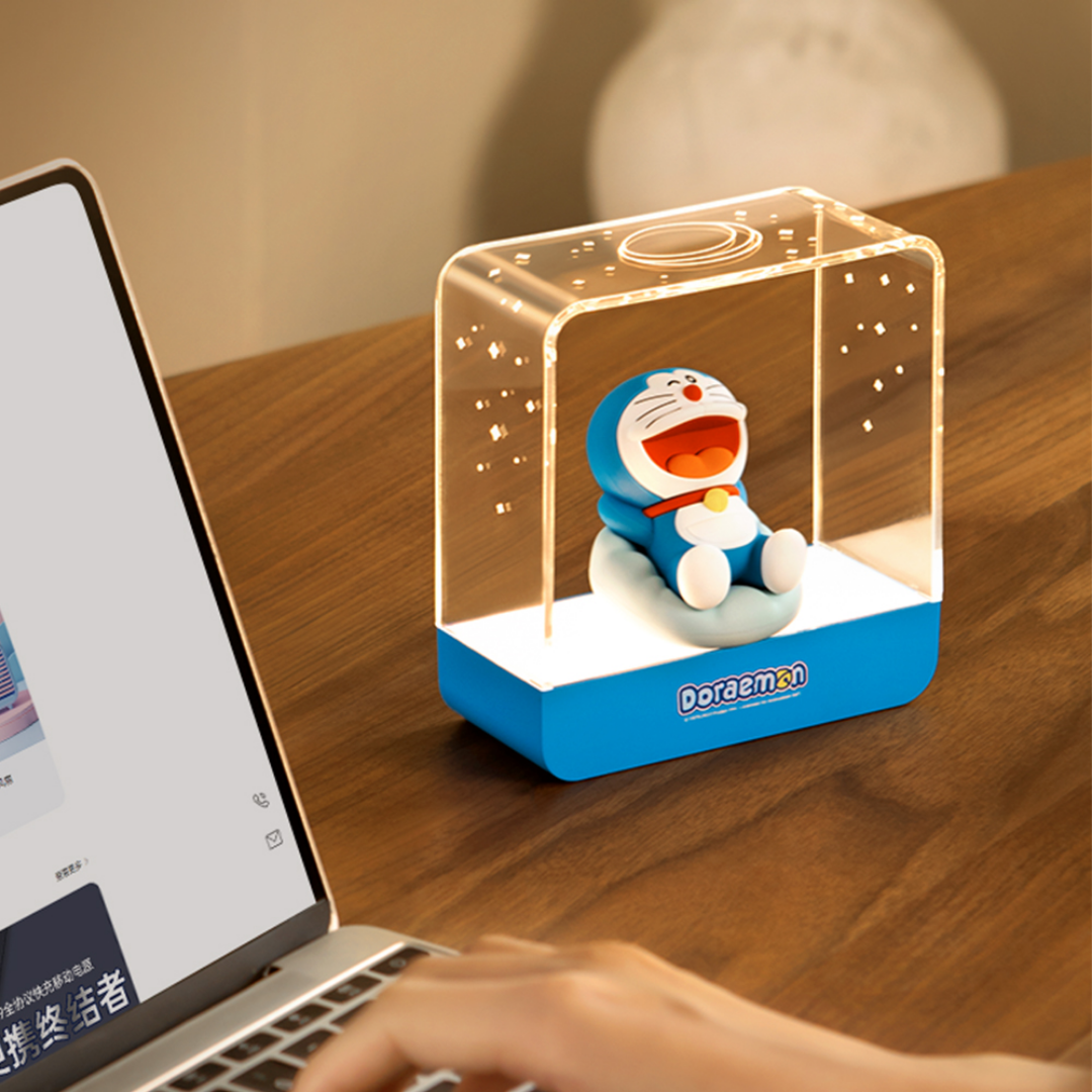 Doraemon Starry Night Light with a smiling Doraemon figure reclining inside a transparent rectangular lamp, projecting warm white star patterns, placed on a wooden desk next to a laptop.