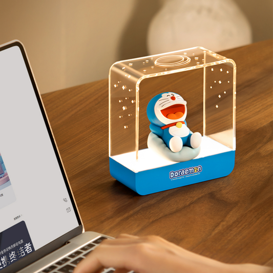 Doraemon Starry Night Light with a smiling Doraemon figure reclining inside a transparent rectangular lamp, projecting warm white star patterns, placed on a wooden desk next to a laptop.
