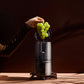 Hurom H310A Slow Juicer black model, BPA-free compact juicer with hand inserting green grapes into hopper, lifestyle action shot with dramatic lighting