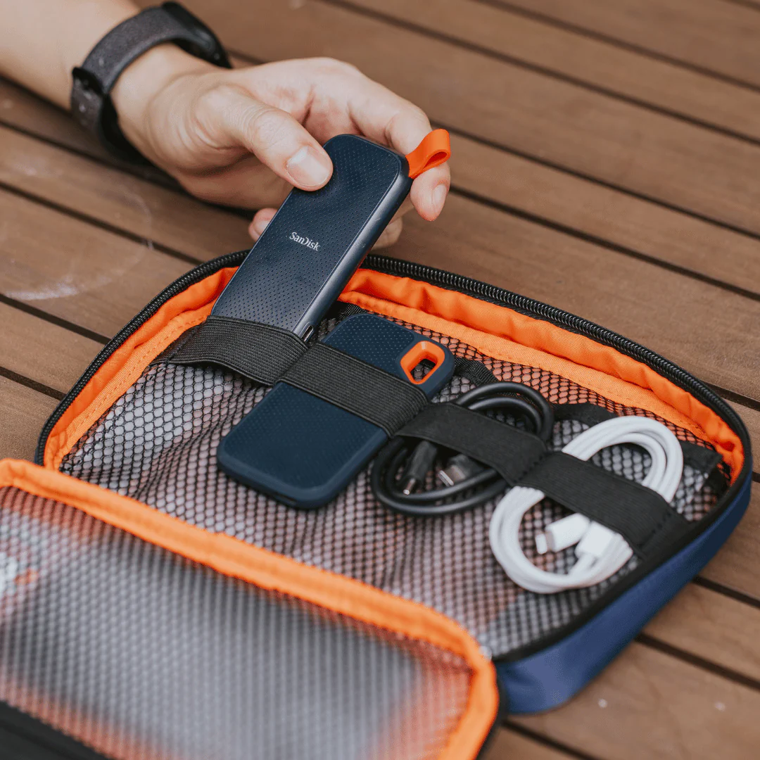Hand organizing portable SanDisk external SSD, charging cables, and accessories in an orange travel tech organizer case on a wooden table.