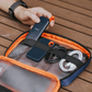 Hand organizing portable SanDisk external SSD, charging cables, and accessories in an orange travel tech organizer case on a wooden table.
