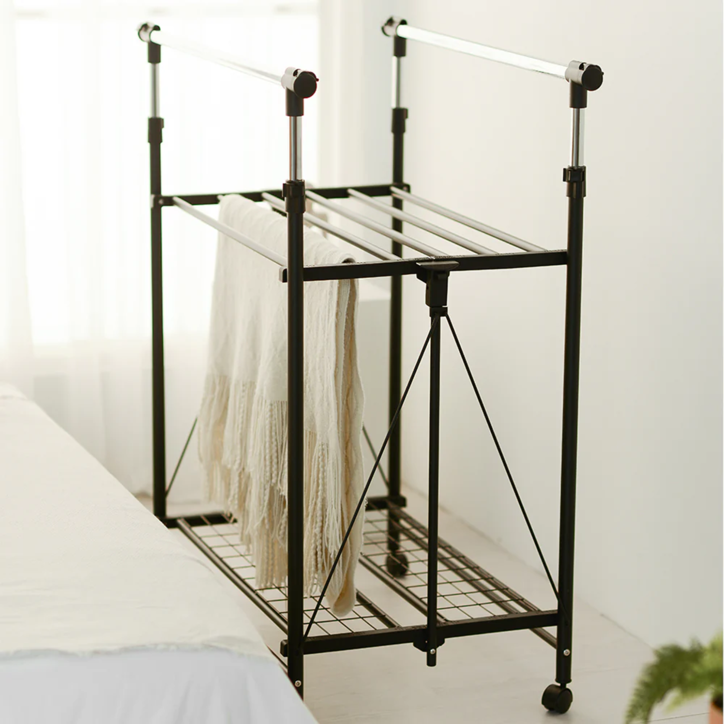 RENE Ocho expandable laundry drying rack in a bright bedroom with a beige blanket hanging over the middle bars and a lower wire shelf for extra drying space.