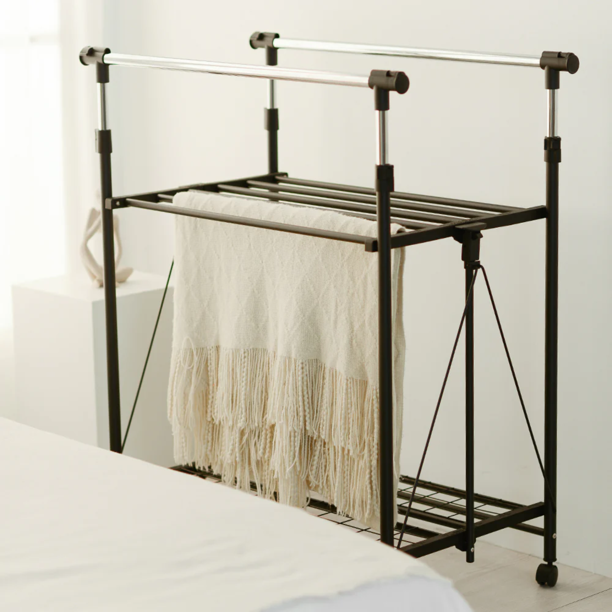 RENE Ocho expandable laundry drying rack in a bedroom holding a beige blanket, showing double bars and lower shelf for space-saving indoor clothes drying.