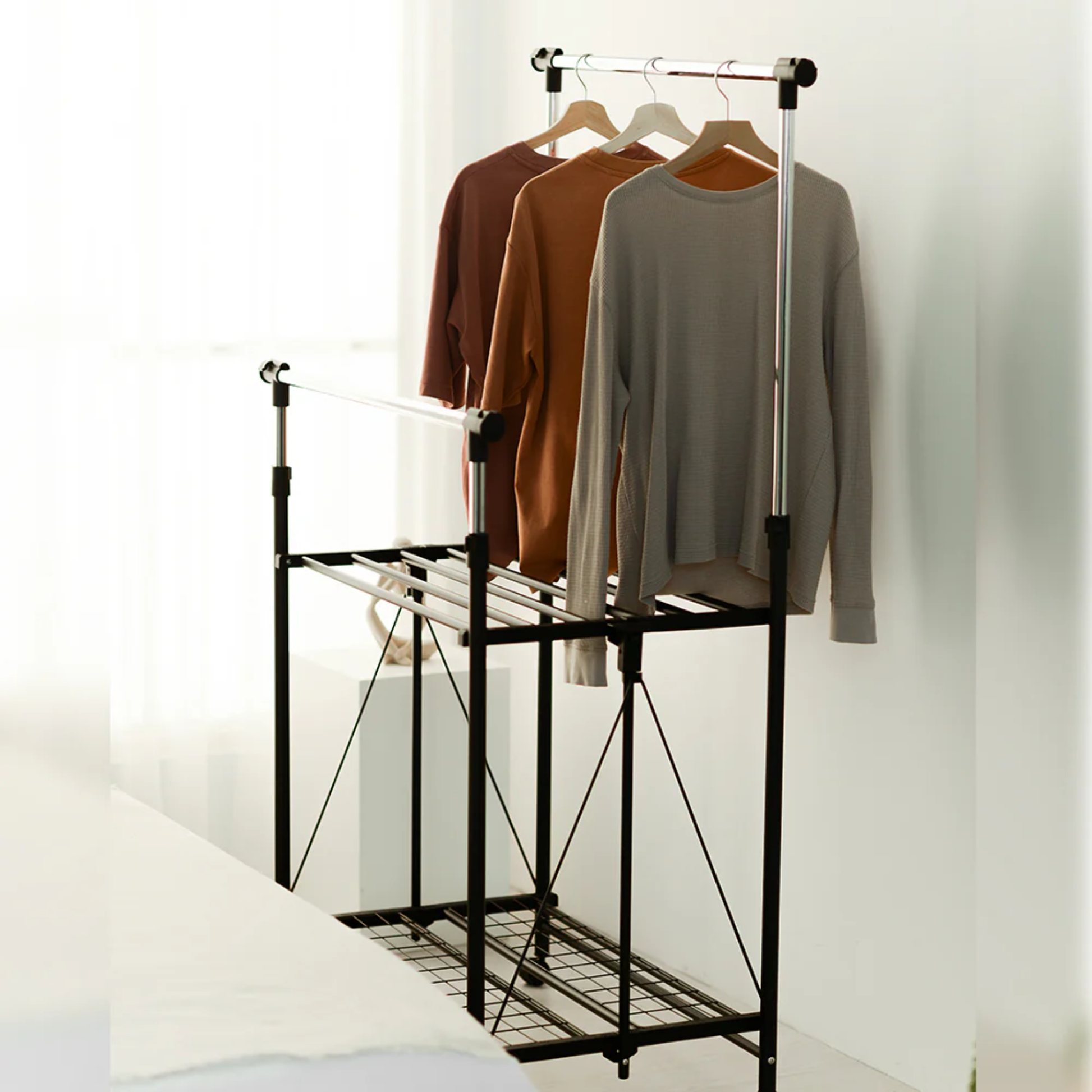 Side view of RENE Ocho expandable laundry drying rack in a bedroom with three long-sleeve tops hanging on the upper bar and a lower wire shelf for extra drying space.
