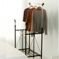 Side view of RENE Ocho expandable laundry drying rack in a bedroom with three long-sleeve tops hanging on the upper bar and a lower wire shelf for extra drying space.