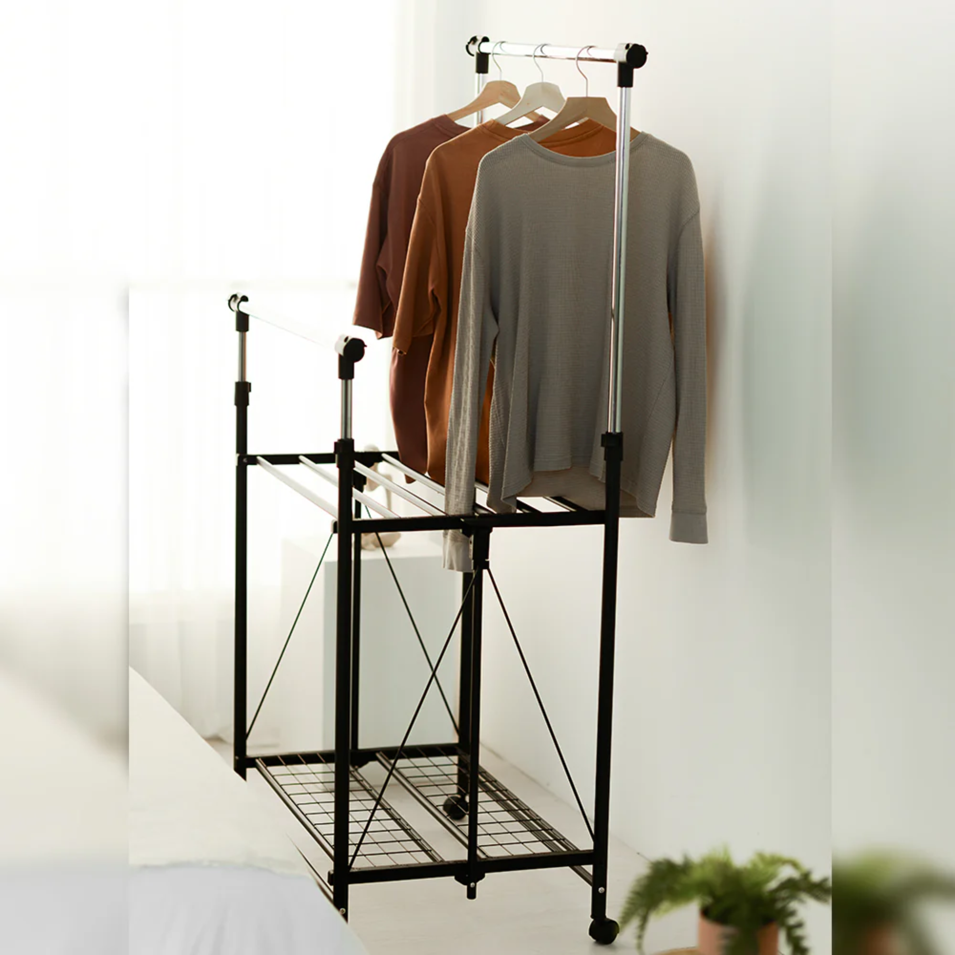 RENE Ocho expandable laundry drying rack in a bedroom with long-sleeve tops hanging on the upper bar and a lower wire shelf for extra clothes drying space.