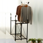 RENE Ocho expandable laundry drying rack in a bedroom with long-sleeve tops hanging on the upper bar and a lower wire shelf for extra clothes drying space.