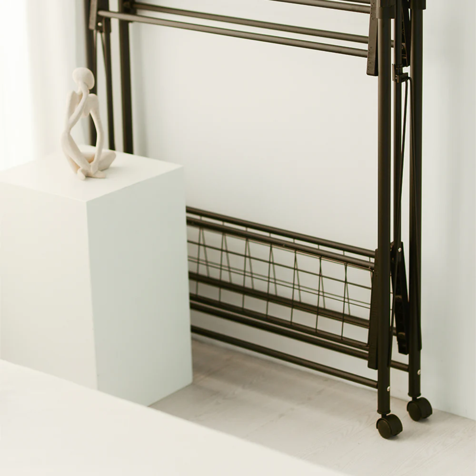 Side view of RENE Ocho foldable laundry drying rack stored upright on wheels beside a white pedestal, highlighting slim, space-saving indoor storage design