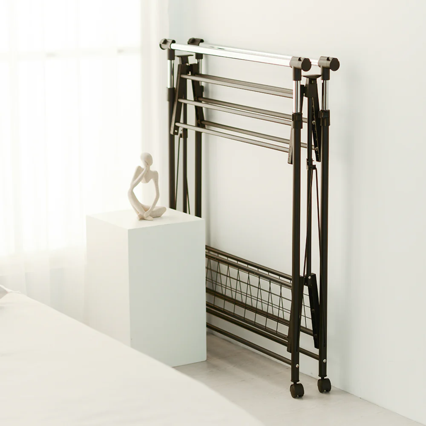 RENE Ocho foldable laundry drying rack stored upright on wheels beside a bed, showing slim space-saving indoor storage in a modern bedroom.