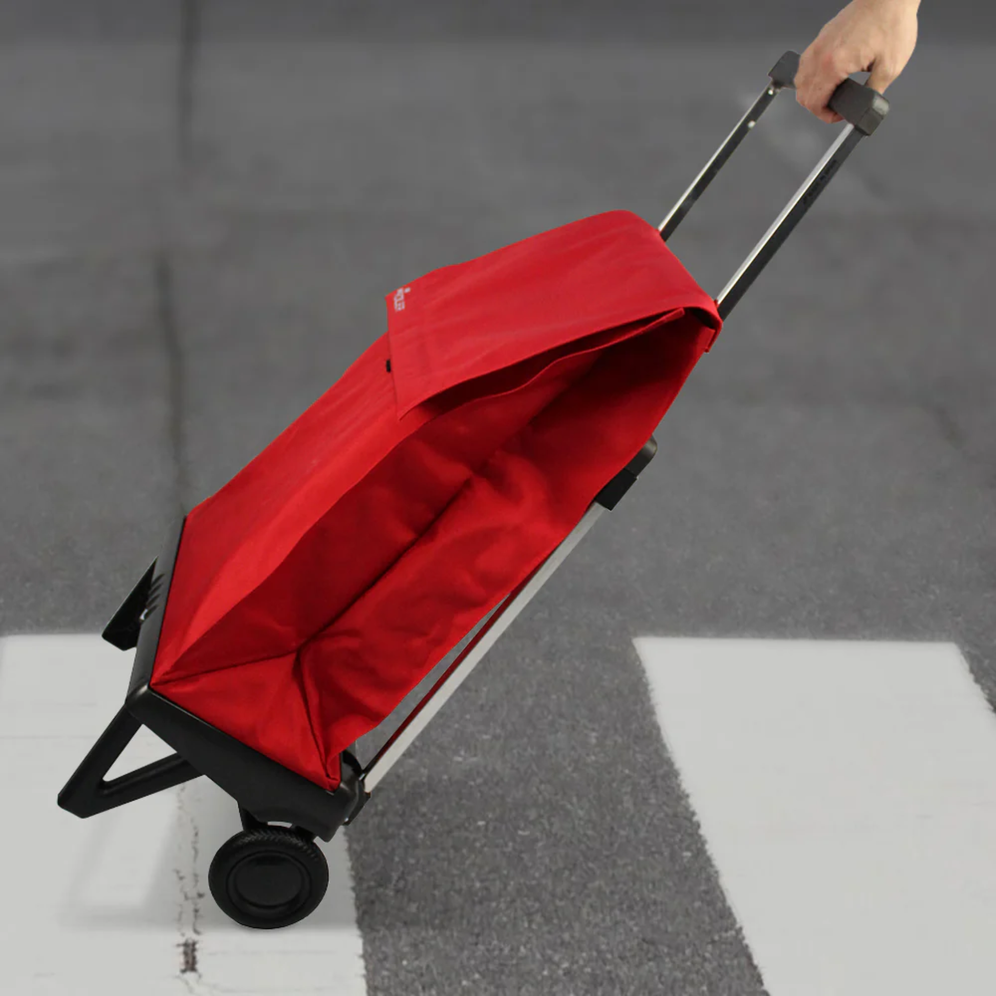 Person pulling a red Rolser Plegamatic foldable shopping trolley with telescopic handle and 2 wheels across a street, showing smooth rolling design.