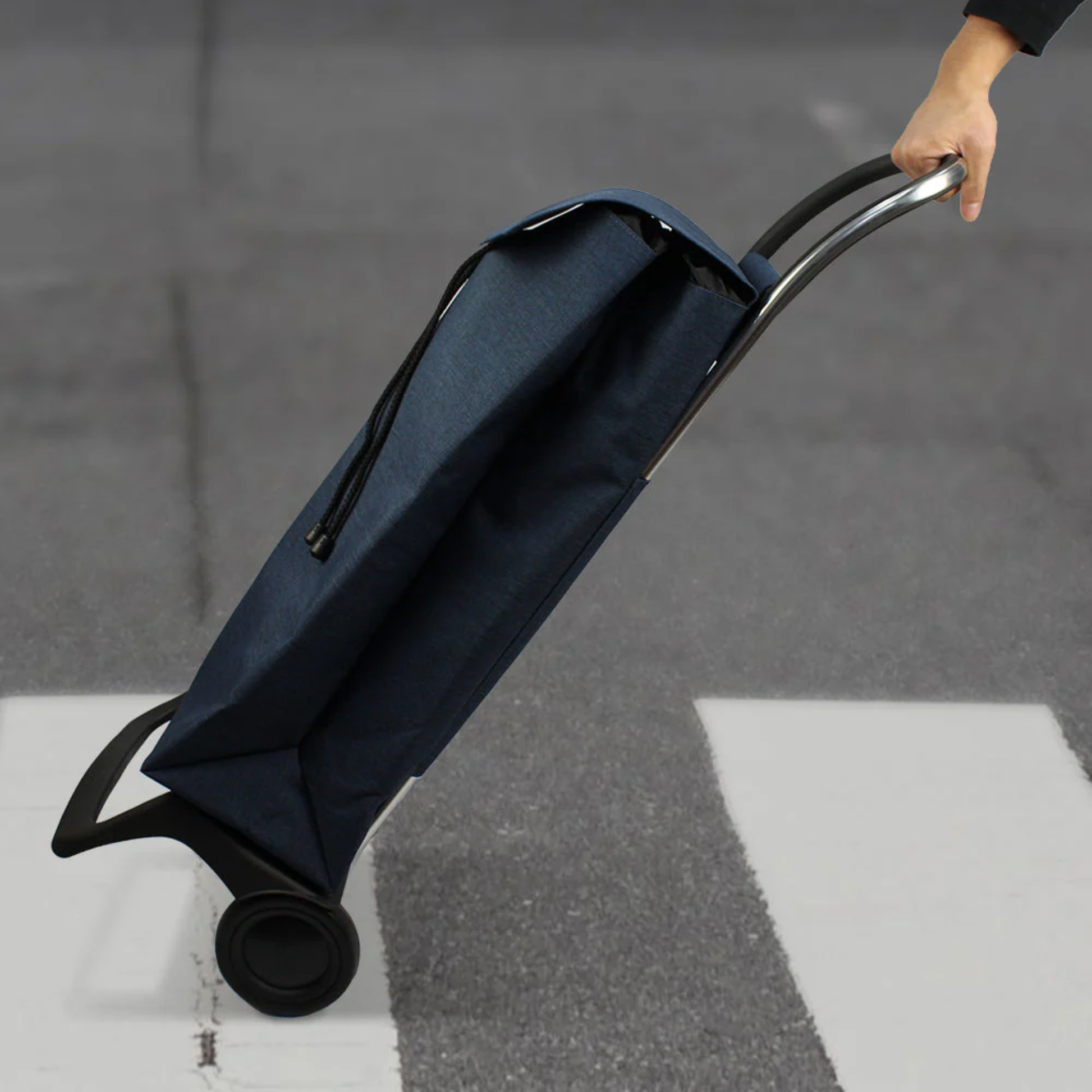 Person pulling a blue Rolser tweed foldable grocery shopping trolley with 2 wheels across a street, showing comfortable handle and smooth rolling.