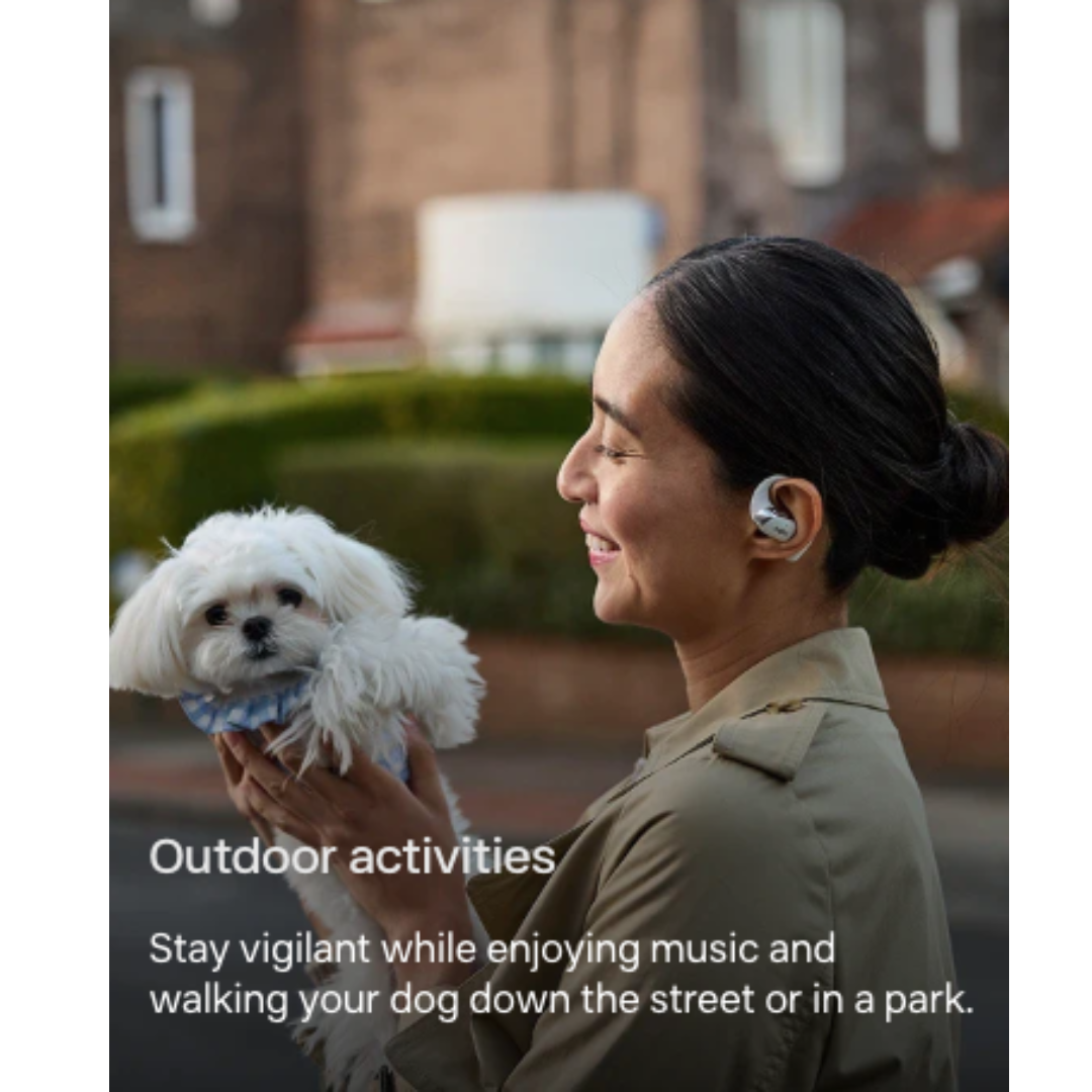 Person wearing Shokz open-ear wireless earbuds walking a small white dog outdoors and enjoying music