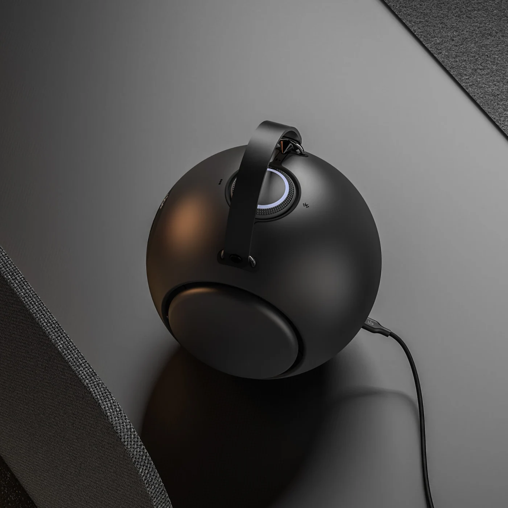 UB+ DB1 Doublebass portable speaker in metallic grey with black carrying handle, shown charging on a modern two-tone desk.